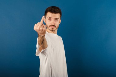 Caucasian man wearing white tee posing isolated over blue background showing middle finger doing fuck you bad expression.