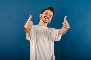Mavi arka planda izole edilmiş tişört giyen mutlu bir adam büyük başarı için el hareketi yapıyor. İki elini de havaya sok..