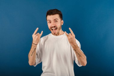 Beyaz tişört giyen yakışıklı beyaz adam mavi arka planda izole edilmiş bir şekilde poz veriyor ve elleri yukarıda çılgın bir ifadeyle rock sembolü yapıyor. Müzik yıldızı. Ağır konsept.
