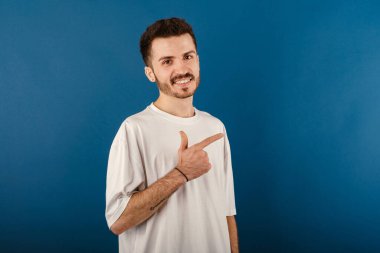 Neşeli beyaz adam mavi arka planda izole bir şekilde poz veriyor işaret parmağıyla gülümsüyor ve kameraya bakıyor..