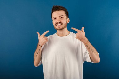 Mavi arka planda izole edilmiş beyaz tişört giyen mutlu genç adam parmakları, dişleri ve ağzıyla gösteriş yapıyor. Diş sağlığı konsepti.