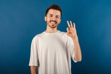 Beyaz tişört giyen beyaz bir adam mavi arka planda izole edilmiş şekilde poz veriyor. Parmaklarıyla iyi bir işaret gösteriyor. İyiliğin sembolü, üst, en iyi, şampiyon, tatmin olmuş ve gururlu..