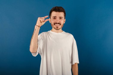 Yakışıklı, günlük kıyafetler giyen, mavi arka planda izole edilmiş bir şekilde poz veren ve parmaklarıyla ölçü alan genç bir adam. Acıyarak küçük boy uzunluğu gösteriyor.