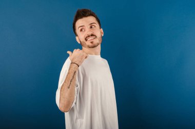 Mavi arka planda izole edilmiş beyaz tişört giyen mutlu genç adam gülümsüyor ve başparmağıyla yan tarafa bakıyor..