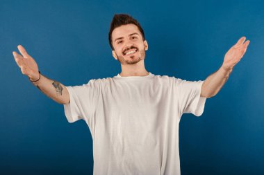 Mavi arka planda izole edilmiş beyaz tişört giyen mutlu genç adam sarılmak için kollarını açmış gülümsüyor. Mutluluğu kucaklayan neşeli bir ifade..