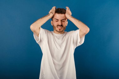 Beyaz tişört giyen bir adamın portresi mavi arka plan üzerinde izole edilmiş bir şekilde duruyor. Başında ağrı var çünkü stres var. Migren ağrısı..