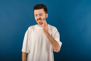 Beyaz tişört giyen beyaz bir adam mavi arka planda yalnız poz veriyor acı çekiyor, yanağını eliyle tutuyor ve kötü diş ağrısından muzdarip. Acı verici bir ifade..