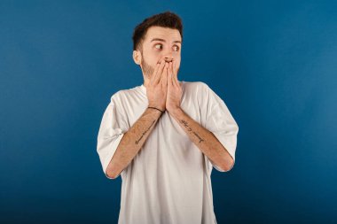 Beyaz tişört giyen yakışıklı beyaz adam mavi arka planda yalnız poz veriyor. Şok olmuş bir şekilde ağzıyla ağzını kapatan bir fotokopi odasına bakıyor..