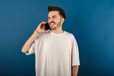 Tişört giyen neşeli genç bir adam mavi arka planda yalnız poz veriyor akıllı telefondan konuşuyor ve gülümsüyor. Telefon görüşmeleri sırasında iyi haberler alınıyor.