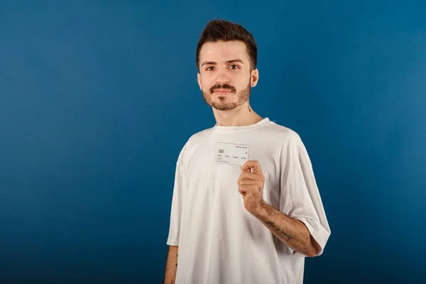 Mavi arka planda yalnız poz veren mutlu sıradan bir adam kameraya bakarken plastik kredi kartı ya da banka kartı taşıyor..