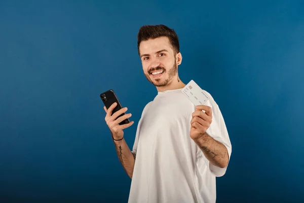 stock image Portrait of young caucasian man wearing t-shirt posing isolated over blue background holding credit or debit card and smartphone in his hands, free space.