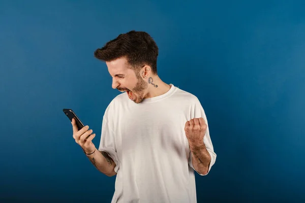 stock image Handsome young man wearing casual clothes posing isolated over blue background being happy winning a bet in online sport gambling application on his mobile phone.