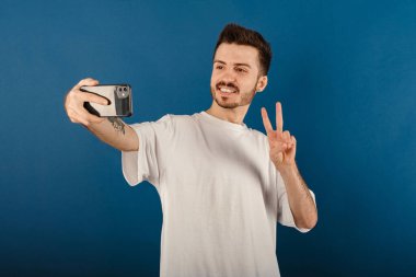 Neşeli beyaz adam mavi arka planda izole bir şekilde poz veriyor zafer işareti yapıyor ve selfie çekerken gülümsüyor. Video çağrısı veya sosyal medya kavramları.
