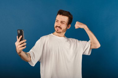 Beyaz tişört giyen neşeli bir adamın portresi mavi arka planda izole edilmiş şekilde poz veriyor, selfie çekiyor, güçlü kollar gösteriyor, kaslarını esnetiyor ve fotoğraf için poz veriyor..