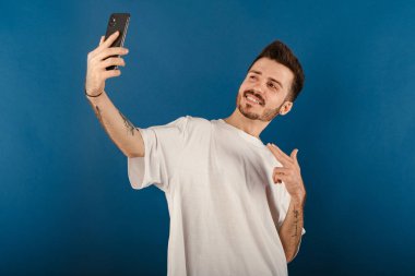 Yakışıklı genç adam mavi arka planda izole edilmiş bir şekilde poz veriyor cep telefonuna işaret ediyor ve gülümserek selfie çekiyor. İletişim kavramları.