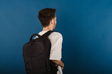 Beyaz tişört giyen beyaz bir adam mavi arka plan öğrencisinin üzerinde yalnız poz veriyor ve sırt çantası takıyor. Arka plan fotoğrafı.