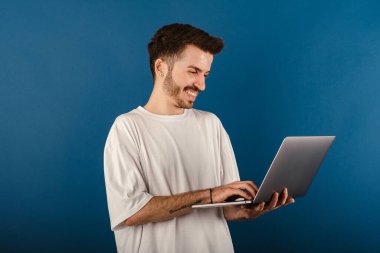 Neşeli beyaz bir adam mavi arka plan üzerinde izole edilmiş bir şekilde poz veriyor ve dizüstü bilgisayarını mutlu ve havalı bir gülümsemeyle gülümsüyor. Dişler gösteriliyor.