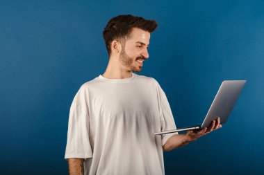 Mavi arka plan üzerinde izole edilmiş beyaz tişört giyen mutlu genç adam ayakta duruyor ve yüzünde mutlu ve havalı bir gülümsemeyle gülümseyen dizüstü bilgisayar kullanıyor. Dişler gösteriliyor.