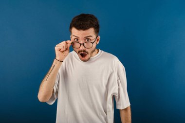 Yakışıklı genç adam mavi arka planda izole edilmiş bir şekilde poz veriyor şaşkınlık ve şaşkınlık içinde bir ifade, korku ve heyecan dolu bir yüz. Gözlüklere dokunmak.