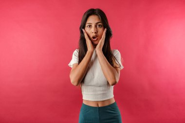 Koyu renk saçlı genç bir kadın kırmızı arka planda izole edilmiş kırbaç takıyor. Ağzı açık kameraya bakıp çenesine elleriyle dokunduğuna inanamıyor. Vay canına..