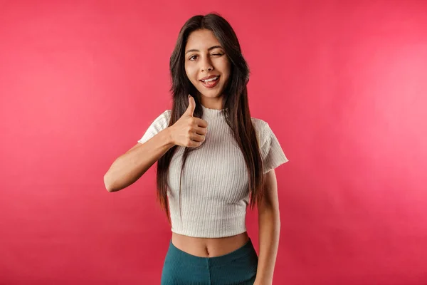 Beautiful brunette woman wearing white ribbed crop isolated over red background smiling and raising thumb up and winking. Thumbs up sign gesture looking at you camera.