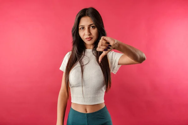 Young caucasian woman wearing casual top isolated over red background not believing the lies, looks at the camera and gives the thumbs down sign. Don\'t tell lies. I didn\'t believe it.