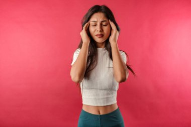 Kaburgalı mahsul giyen genç, siyah saçlı kadın kırmızı arka planda izole edilmiş ve baş ağrısı şikayetiyle şakaklarına masaj yapıyor. İyileşmek istiyor. İnsanlar, duygular, stres ve sağlık hizmetleri kavramı.