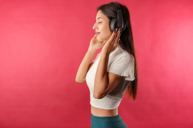 Kırmızılı kırbaç giyen genç, siyah saçlı kadının yan görüntüsü. Kırmızı arka planda izole edilmiş. Kulaklıkla müzik dinlemek, dans etmek. Favori müzikle dans etmek çok mutludur. Bundan çok zevk alıyor. Saç savuruyor..