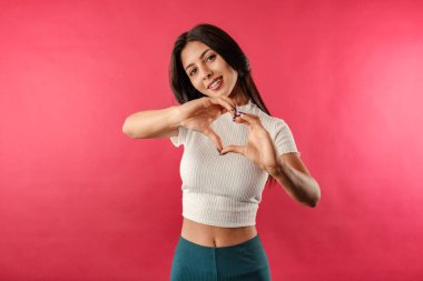 Kırmızı arka planda izole edilmiş beyaz kurdeleli genç bir kadın elleriyle kalp işareti yapıyor ve gülümseyerek kameraya bakıyor. Kalp sembolü ve şekilli eller. Romantik konsept.