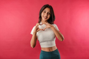 Kırmızı arka planda izole edilmiş, gündelik giyinen mutlu esmer kadın elleriyle kalp sembolü çiziyor. Romantik konsept.