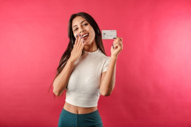 Kırmızılı tarla giyen genç kahverengi saçlı kadının portresi kırmızı arka planda izole edilmiş, elinde kredi kartı var. Üyelik kartı maketi alışveriş parasını internetten gösteriyor..