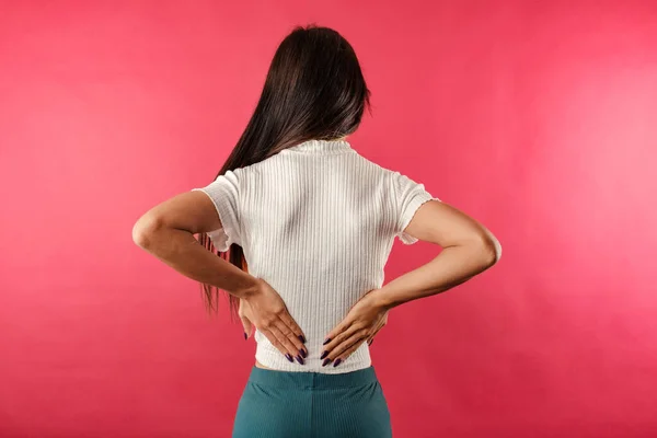 Brown-haired woman wearing white ribbed crop isolated over red background with very severe back pain holds back with hand and shows the pain. View from the back.