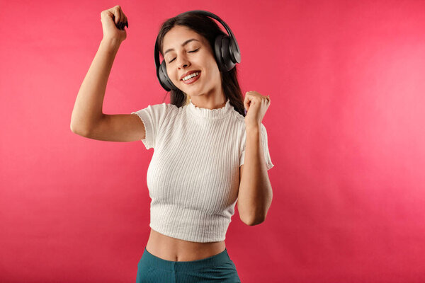 Portrait of young brown-haired woman wearing ribbed crop isolated over red background listen music with headphones, dancing. Dancing to favorite music is very happy. Enjoys it very much.