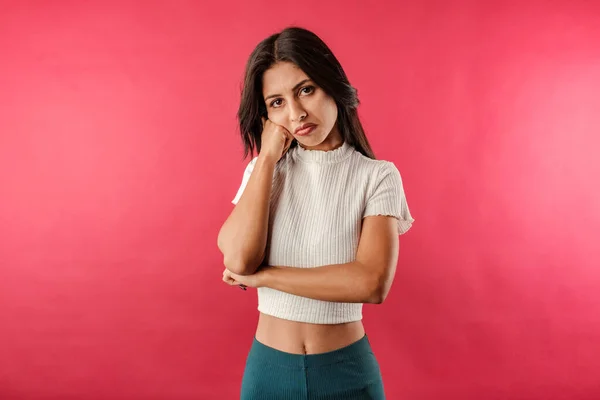 Portrait of young brunette woman wearing white ribbed crop isolated over red background very bored and waits with head resting on the fist. It is so boring to wait, to listen to the lies being told.