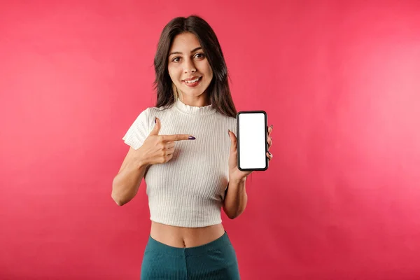 Young dark-haired girl smiling happy wearing ribbed crop isolated over red background smiling, points with index finger to the blank white mockup smart phone screen. Ad or some kind of presentation.
