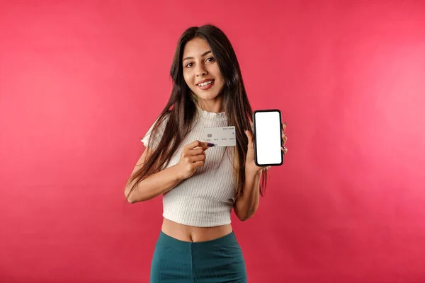 Brown-haired woman isolated over red background holds credit card and phone with blank white mockup screen side by side and shows it to the camera. Looks at the camera with a smile.
