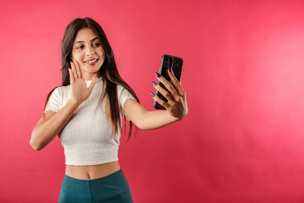 Young caucasian woman wearing casual top isolated over red background making a video call with smartphone and waving palm hand. Talks remotely with boyfriend or family. Laughs. Doing selfie.
