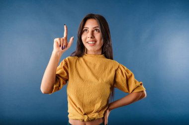Happy young woman wearing yellow t-shirt isolated over blue background looks in amazement as indicates at something upwards. Pointing finger up with successful idea. Exited and happy. number one.