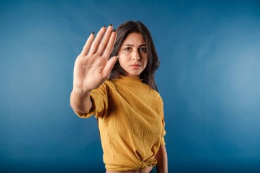 Portrait of brunette woman wearing mustard yellow t-shirt isolated over blue background doing stop sing with palm of the hand. Warning expression with negative and serious gesture on the face.