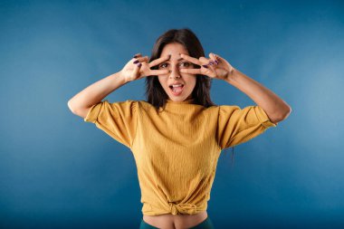 Young dark-haired girl smiling happy wearing yellow top isolated over blue background doing peace symbol with fingers over face, smiling cheerful showing victory. Wow face! Very happy about what hears