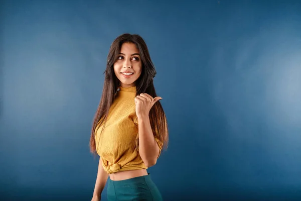Side view of cheerful woman wearing casual top isolated over blue background smiling, looking and pointing to the side with thumb up. Eyes look side empty space. Directing finger banner.