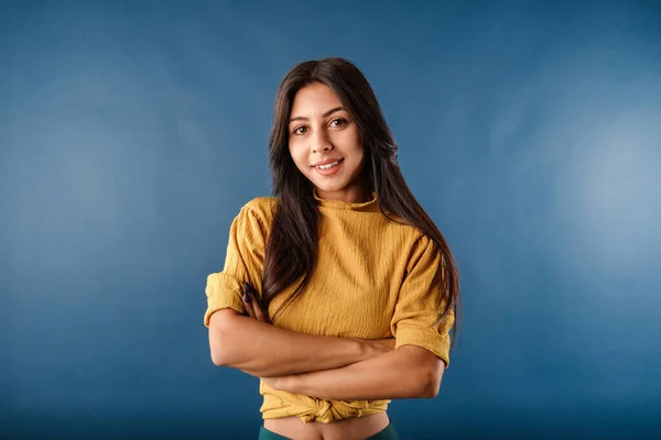 Young woman smiling confident wearing mustard yellow t-shirt isolated over blue background poses with arms crossed, looking at the camera. Looks confident and accomplished.