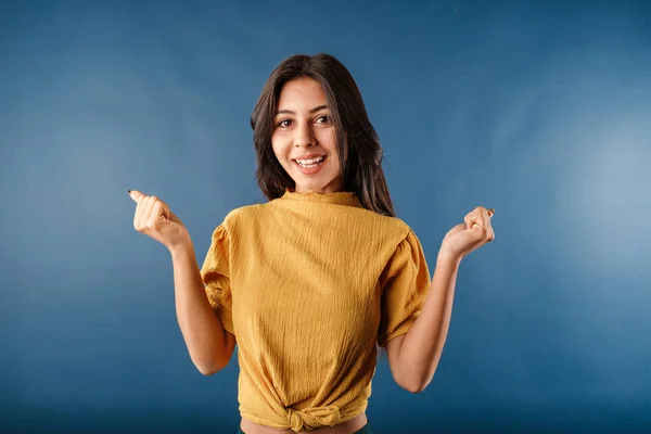 Portrait of young brunette woman isolated over blue background hands in fists shouts for joy and laughs at the camera. Winning and celebrating, triumphing, raising hands up and laughing.