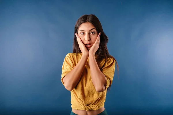 Portrait of cute dark-haired woman wearing casual top isolated over blue background can't believe what hearing and looks at the camera with mouth open and touches chin with hands. Wow face.