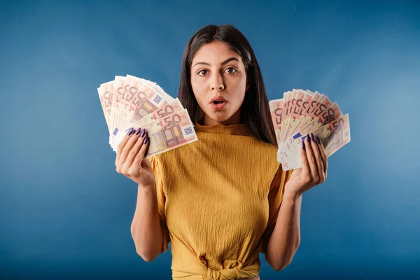 Young dark-haired woman wearing yellow top isolated over blue background holding a wad of cash looks at the camera, scared and amazed and open mouth. Unbelievable. So much money.