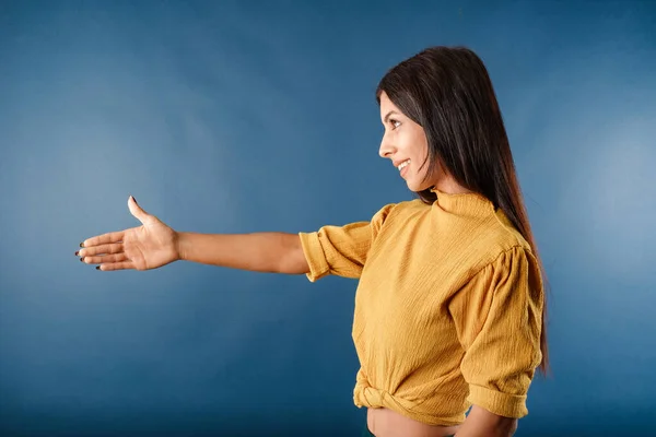 Side view of young brunette woman isolated over blue background giving hand for handshake, greeting with smile. The concept of meeting. Holds out hand to the empty copy space and wants to shake hands.