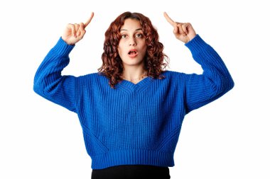 Portrait of cheerful woman wearing pullover sweater standing isolated over white background pointing with two fingers, looking at camera with open mouth. Index fingers pointing up amazed.