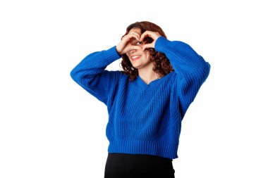 Young beautiful woman isolated over white background doing heart shape with hand and fingers looking through sign. Smiles with love. Makes a heart sign with hands and looks at the camera, at you.