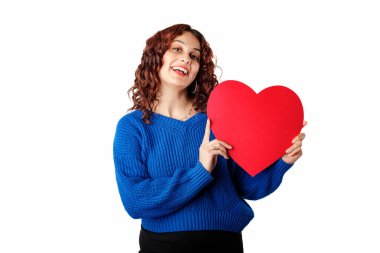 Young beautiful woman wearing blue pullover sweater isolated over white background poses smiling with a cardboard heart shape. Looking at the camera, at you, at lover. It could be a gift.