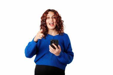 Young girl smiling happy wearing blue sweater isolated over white background holding phone and looking at the camera, at you, with a puzzled expression and pointing at you with index finger.